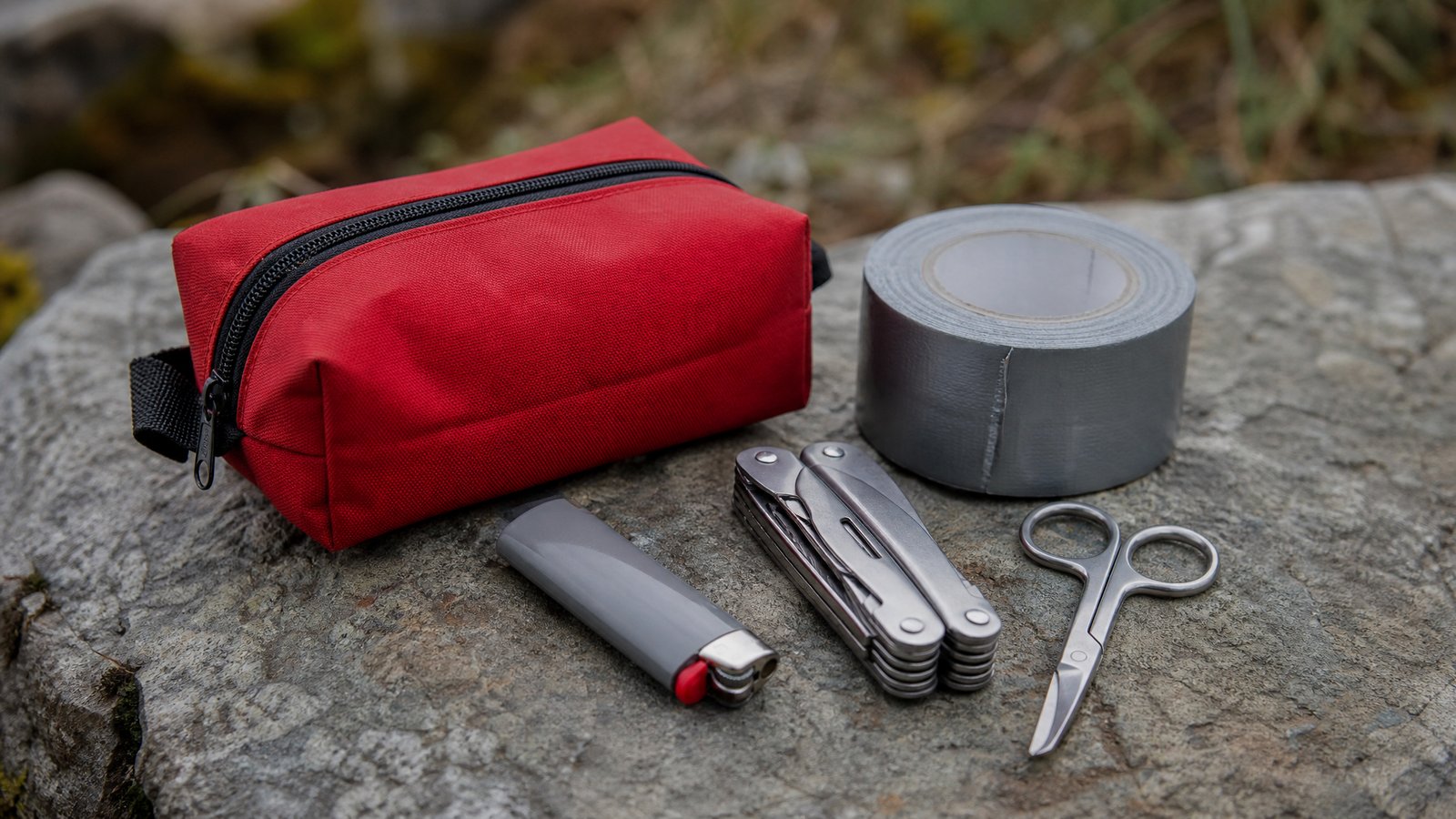 A plain red zippered first aid pouch, a gray lighter, a roll of gray duct tape, a folding multi-tool and small scissors on a flat rock