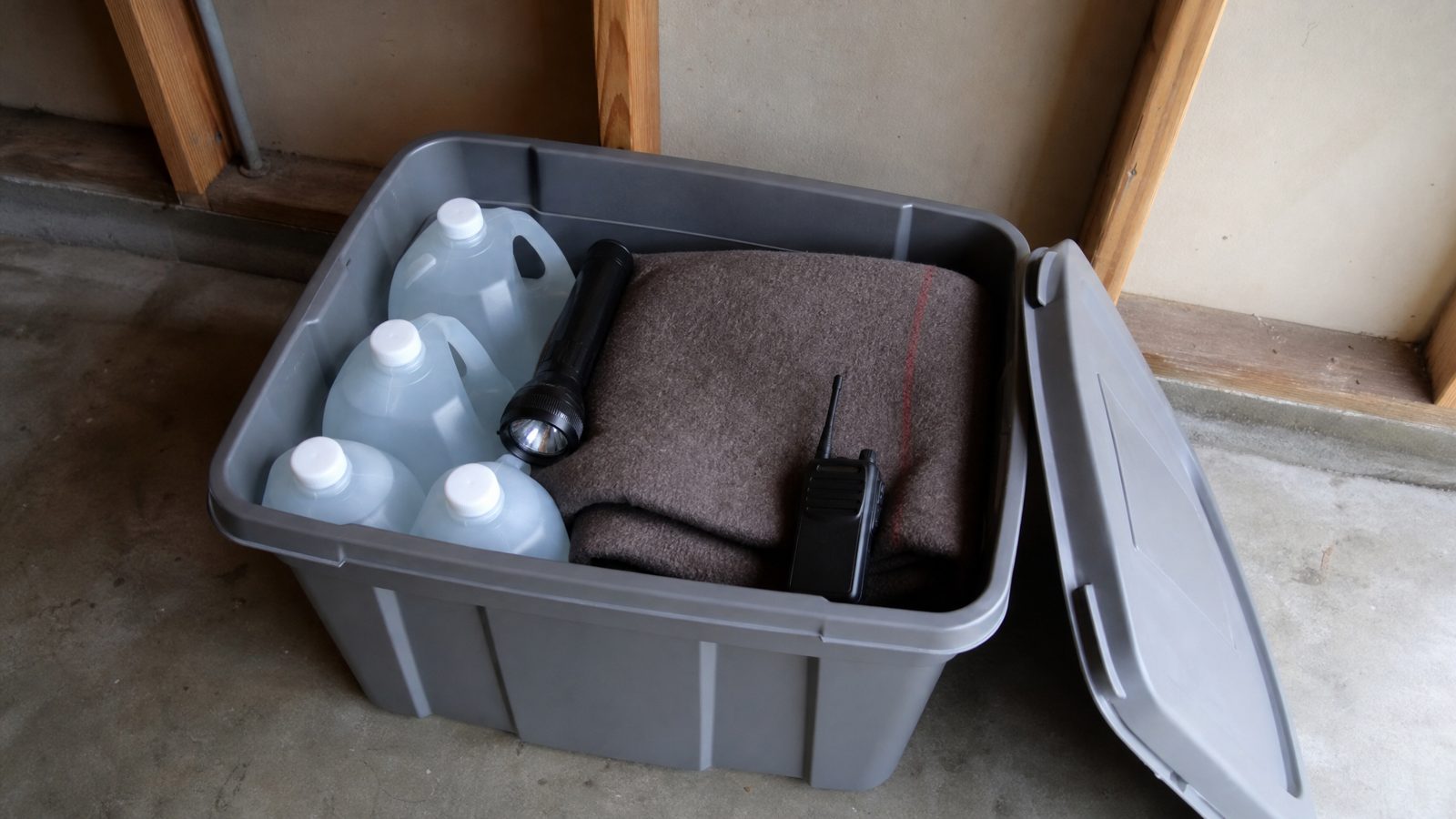 A gray plastic storage bin with the lid off on a garage floor holding water jugs, a black flashlight, a folded wool blanket and a handheld radio