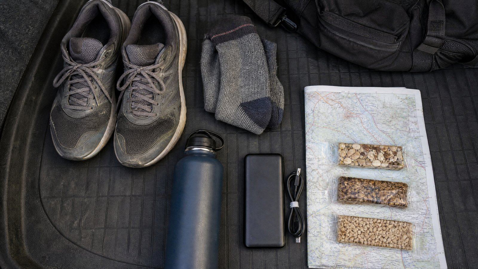 Get-home bag contents laid out on a car trunk liner: worn athletic shoes, folded socks, a water bottle, a battery pack, a folded paper map, and energy bars