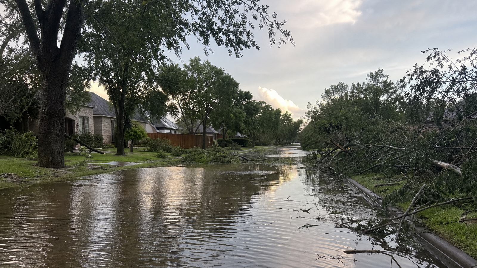 A residential street after a storm with brown standing water over the lower part of the road and fallen tree branches on the grass