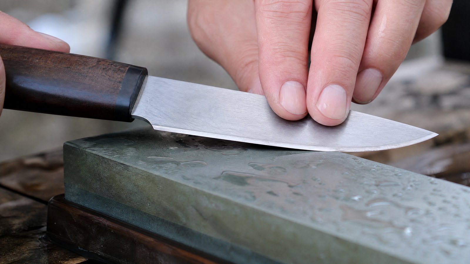 A close-up of a fixed-blade knife edge against a whetstone, hand holding the spine at a low, steady angle