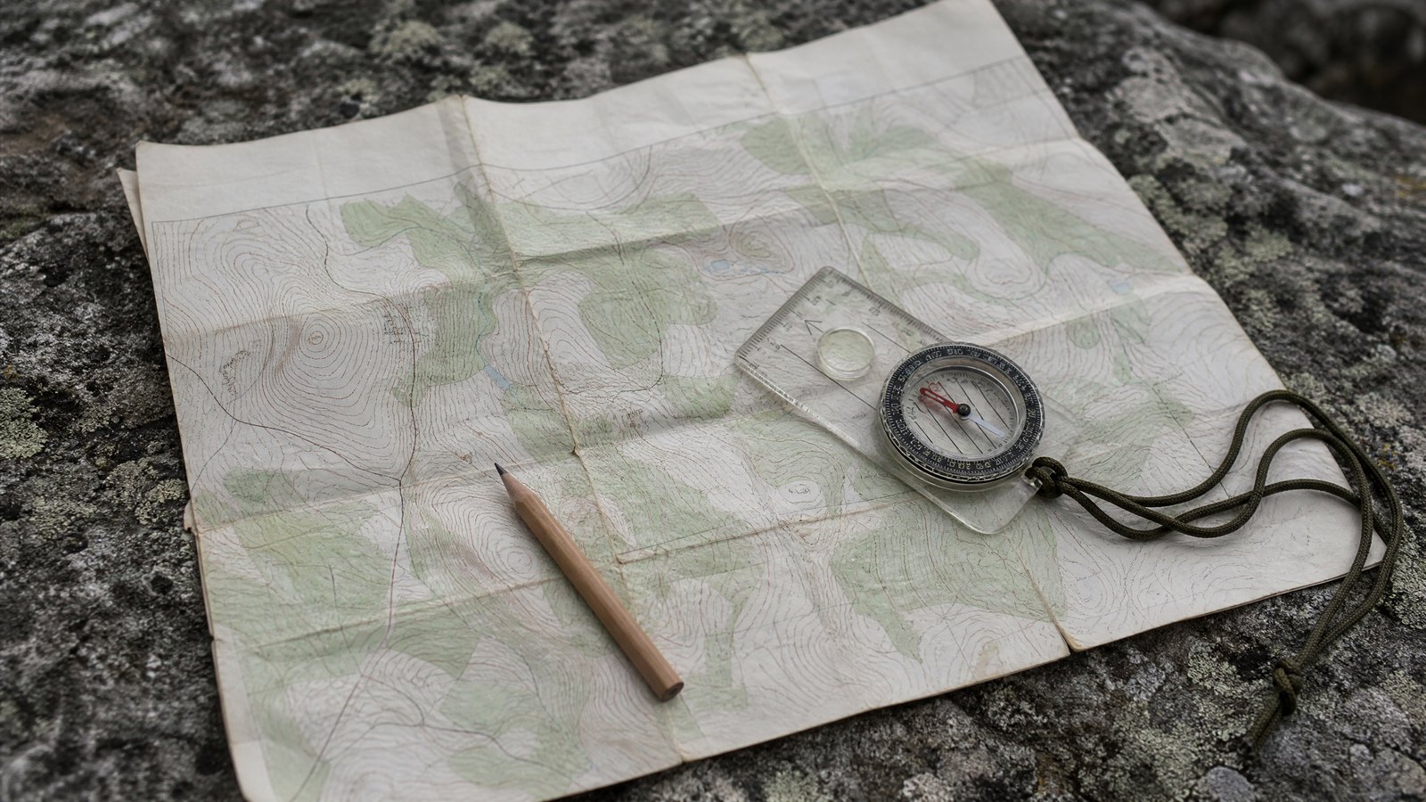 An unfolded paper topographic map on flat rock with a clear baseplate compass, a pencil, and a length of cord laid alongside