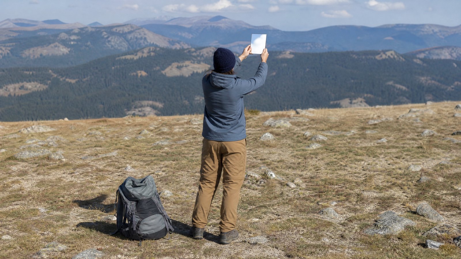 A hiker standing alone in an open alpine clearing angling a small mirror toward the bright sky with hazy ridgelines behind
