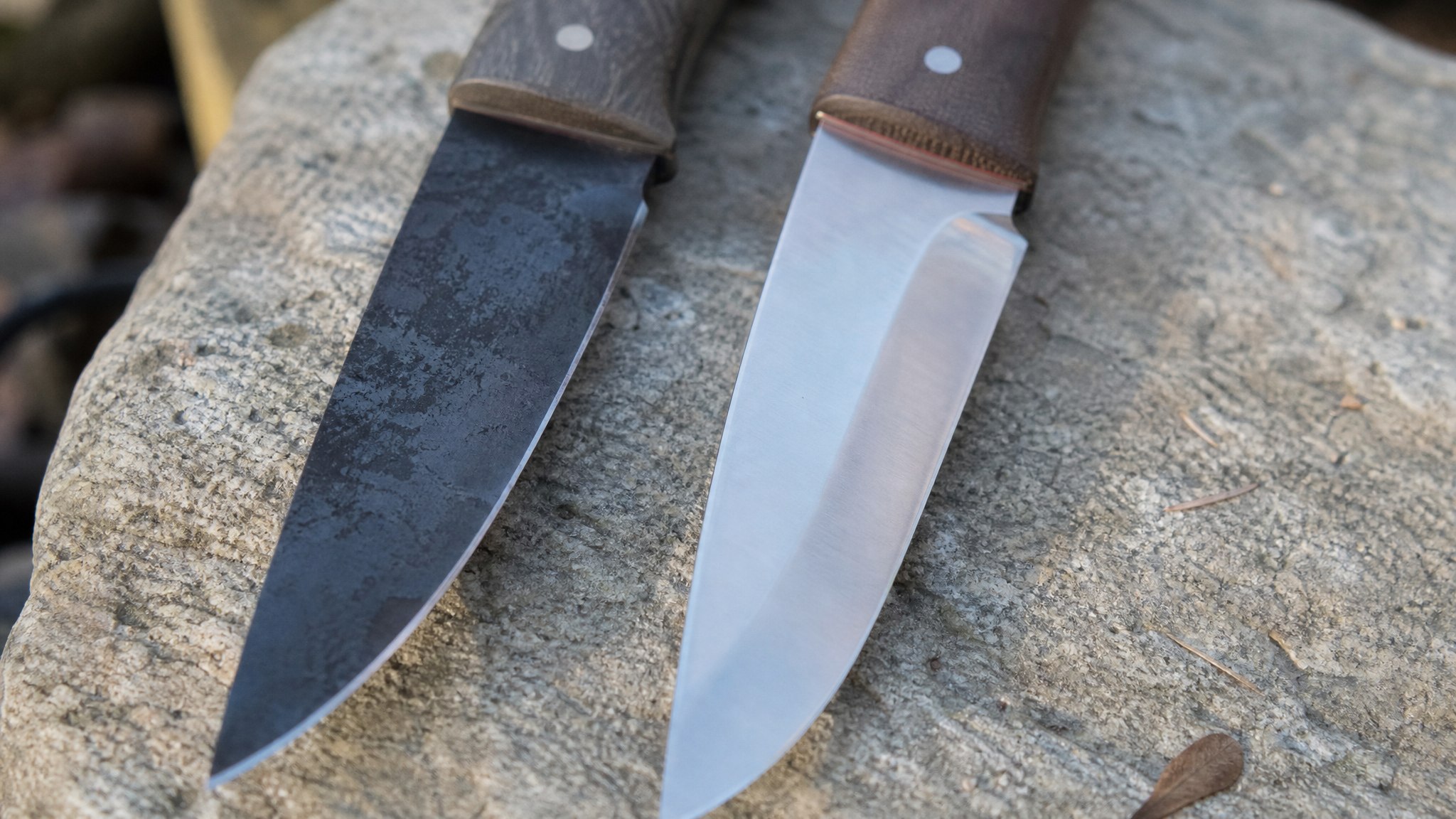 Two knife blades laid side by side on a rock, one showing a dark carbon steel patina and one bright stainless steel finish
