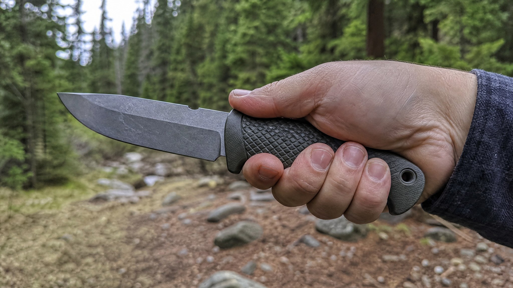 A hand gripping a drop point survival knife with a textured handle, showing the blade length against an outdoor backdrop