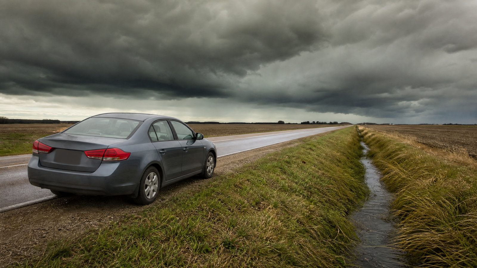 A plain gray sedan parked on the shoulder of a rural two-lane road under a dark green-gray sky, with a drainage ditch and wet grass beside the road