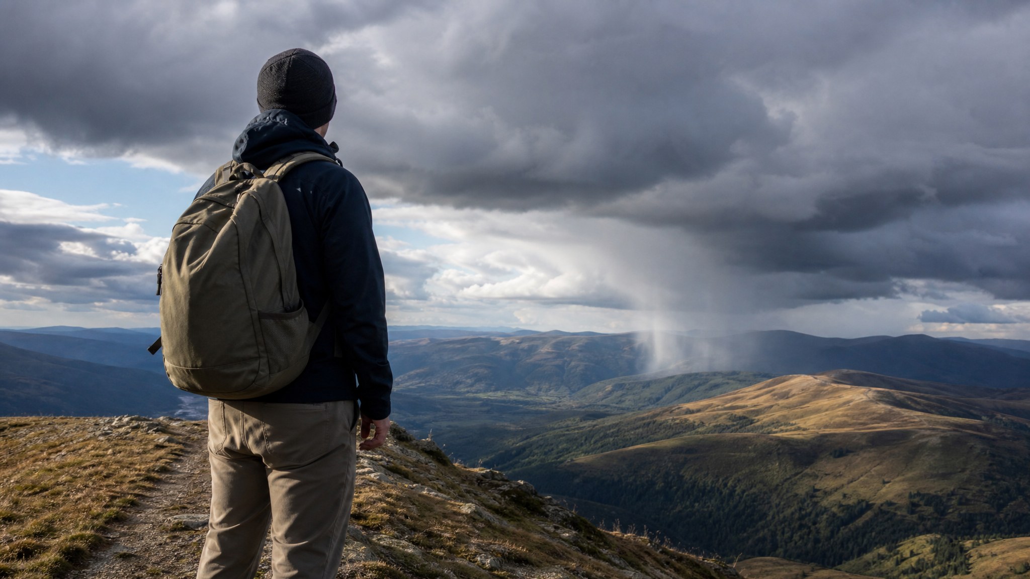 A person standing on a ridgeline trail looking up at a darkening sky with towering clouds building in the distance