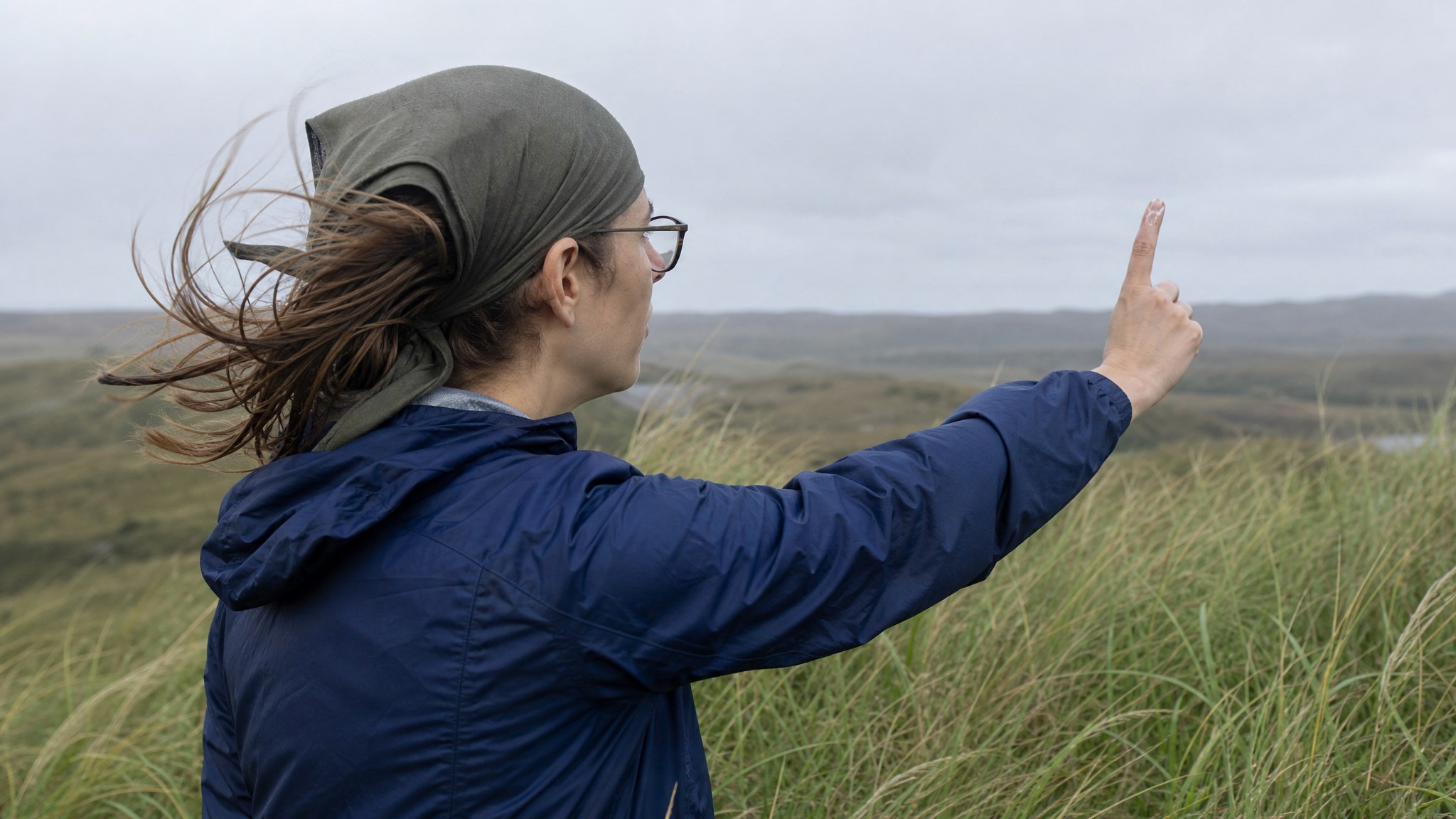 A person outdoors holding up a wetted finger to check wind direction, with tall grass bending sideways in the wind around them
