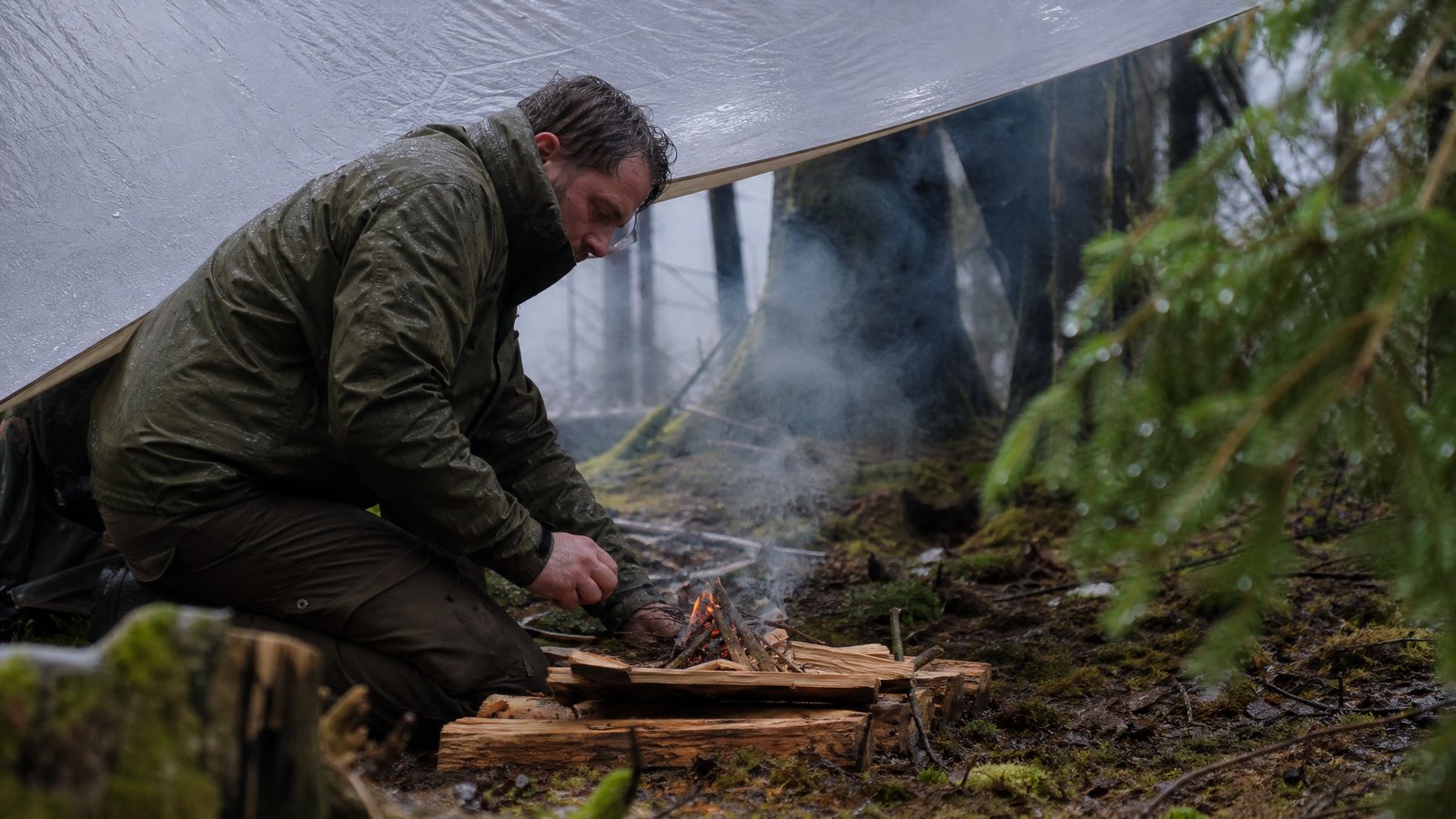 Man crouched under a tarp tending a small campfire on a platform of split wood in a rainy pine forest